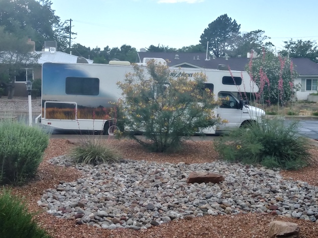 A large RV parked in front of a xeriscaped lawn with green bushes