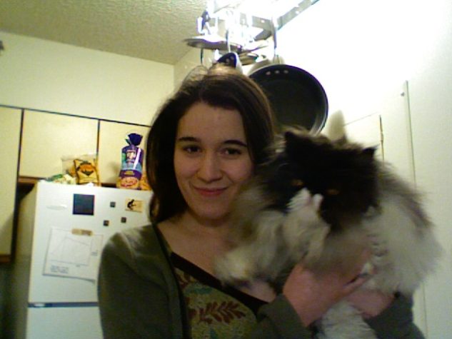 A photo of me, a white woman with long brown hair, wearing a green shirt and sweater, holding a giant fluffy grey and white Persian cat, with a white fridge visible in the background