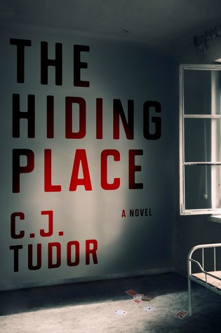 Cover of "The Hiding Place" with title and author C. J. Tudor's name in red text over an empty white room, in shadow, with an open window shutter, edge of a bedframe, and playing cards scattered across the floor. 