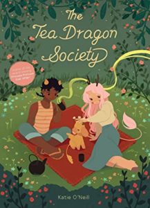 Cover of "The Tea Dragon Society" with the title and author Kay O'Neill's name in script font over the background of an illustration from the comic: 2 human-like characters (one pale with small antlers, pink hair, a pink tail, a blue skirt and beige top, and one dark-skinned person with short horns, flip-flops, blue capris, and an orange striped-tank top. They are on a red blanket in a green meadow, and the person in a tank is drinking tea with scent that wafts over the image, while the person in the skirt is petting a tea dragon, who's yellowish and small.