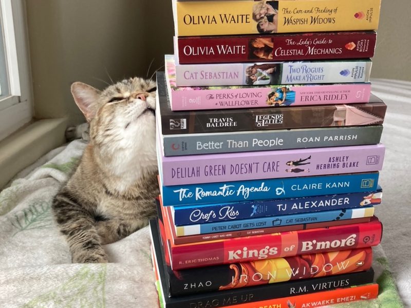 A brown-and-white tabby cat (Tib) rubbing against a tall stack of books (my TBR pile), while laying on a very cat-hair covered blanket on a bed