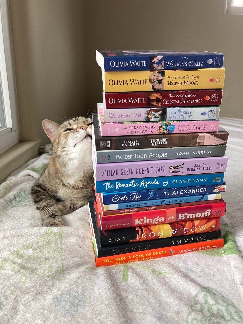 A brown-and-white tabby cat (Tib) rubbing against a tall stack of books (my TBR pile), while laying on a very cat-hair covered blanket on a bed